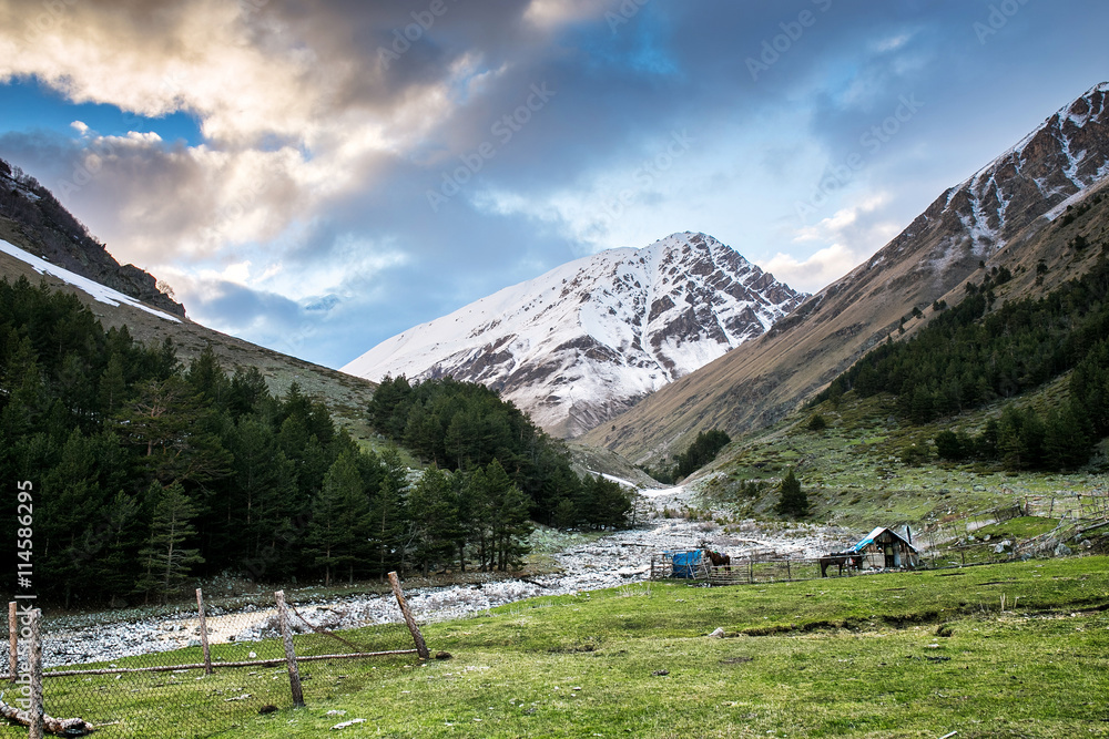 Beautiful mountain landscape of green valley in the Caucasus mountains. In the foreground a small triangular wooden shelter and benches, then fence and house. Russia