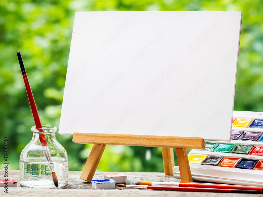 Fototapeta premium Sketchbook on easel, watercolor paints, artistic paintbrushes an