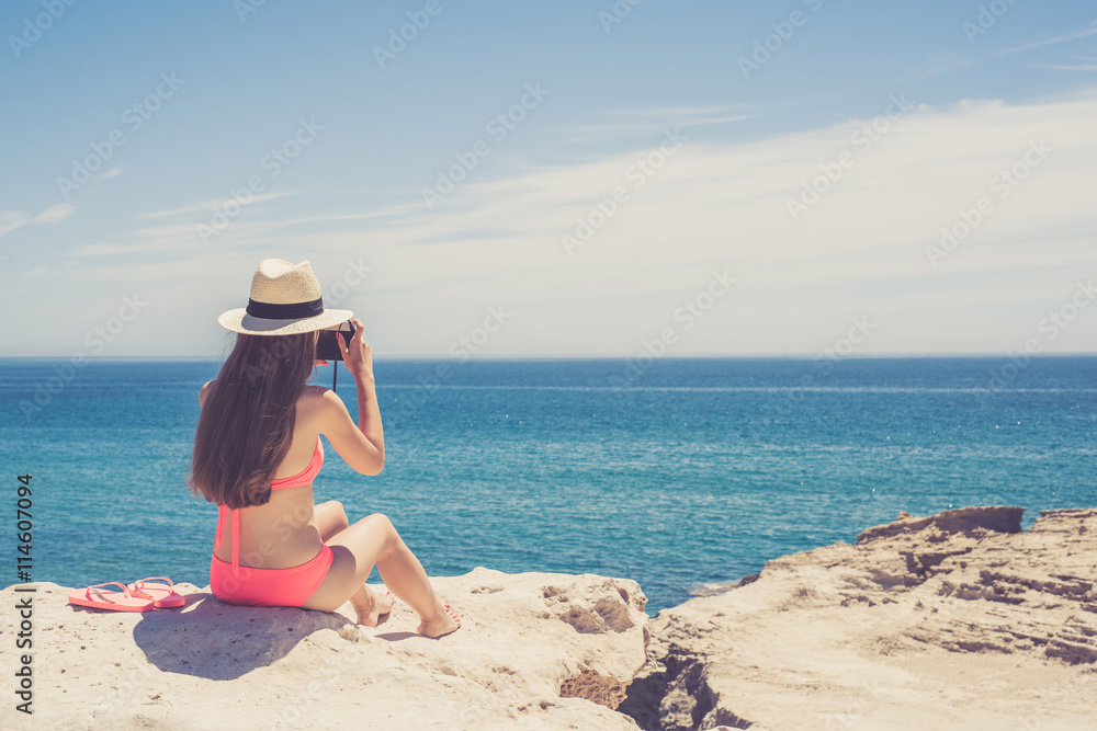 Young woman with long hair sitting on the cliff and taking photo of beautiful exotic sea