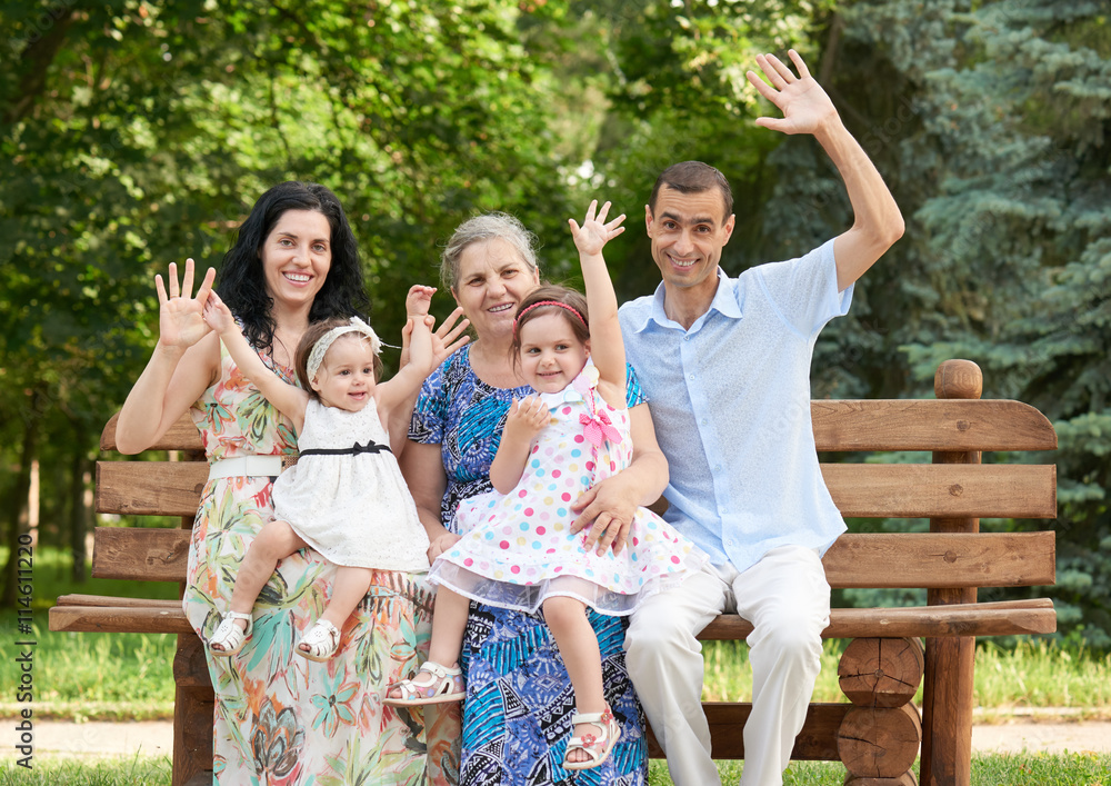 Naklejka premium big family sit on wooden bench in city park and waving, summer season, child, parent and grandmother, group of five people
