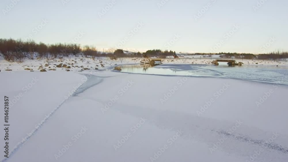 Low altitude aerial from a frozen lake in winter with snow on the ground, people riding horses over a bridge in the background Low altitude aerial from a frozen lake in winter with snow on the ground, people riding horses over a bridge in the background