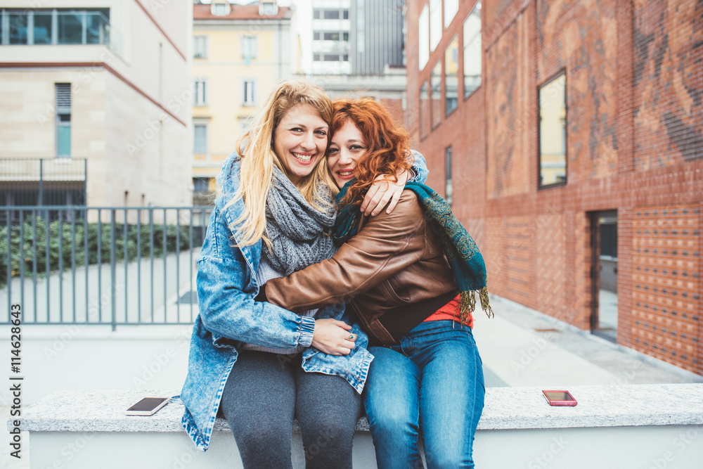 Two friends redhead and blonde girl in the street of the city hugging , looking in camera, smiling –friendship, happiness concept Two friends redhead and blonde girl in the street of the city hugging , looking in camera, smiling –friendship, happiness concept