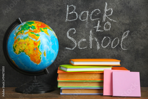 wooden plaque with the inscription "back to school" near the globe on a stack of notebooks on a brown wooden table on blackboard background with inscription "back to school"