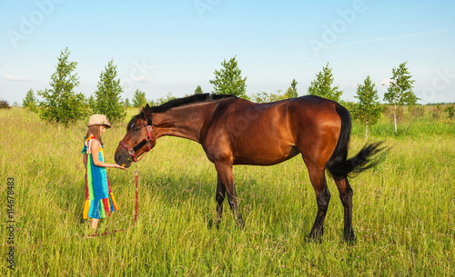 Little girl in bright hat and sundress feeding a horse on the lawn
