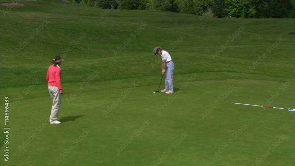 Woman golfer putting as companion helps line up shot, ball rolls short of hole Woman golfer putting as companion helps line up shot, ball rolls short of hole