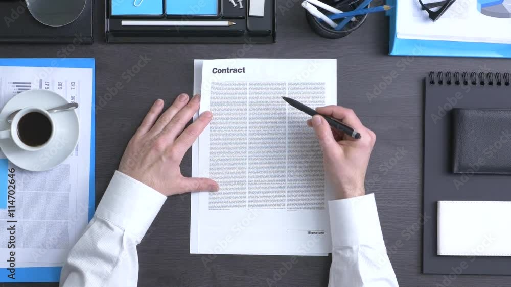 Businessman working at office desk, he is reading and signing a contract, top view Businessman working at office desk, he is reading and signing a contract, top view
