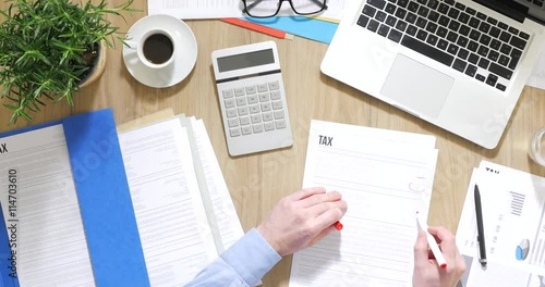 Businessman working at office desk, he is checking tax forms and using a calculator, top view