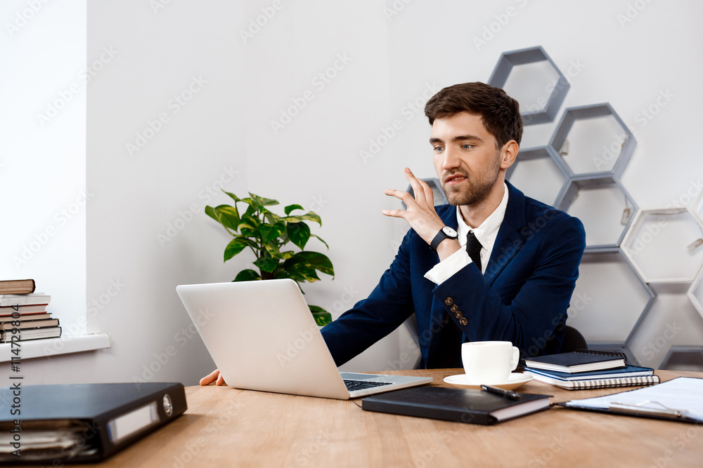 Annoyed young businessman sitting at laptop, office background. Annoyed young businessman sitting at laptop, office background.