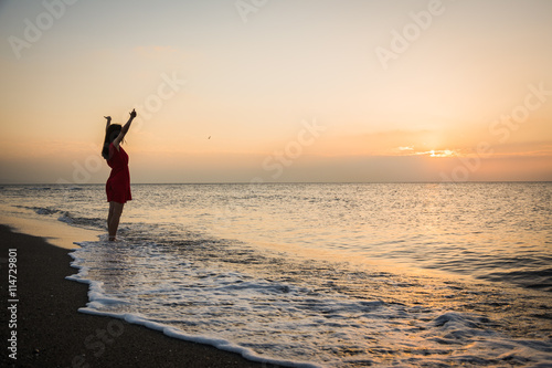 Girl in a red dress at sunrise on a golden beach watching the sea silhouette
