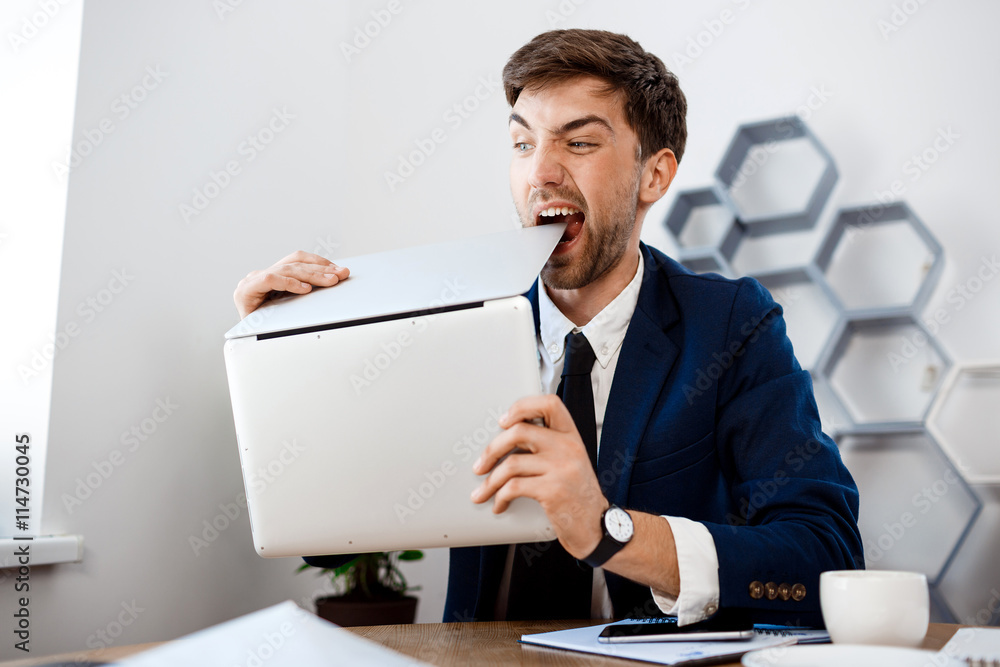 Angry young businessman gnawing laptop, office background. Angry young businessman gnawing laptop, office background.