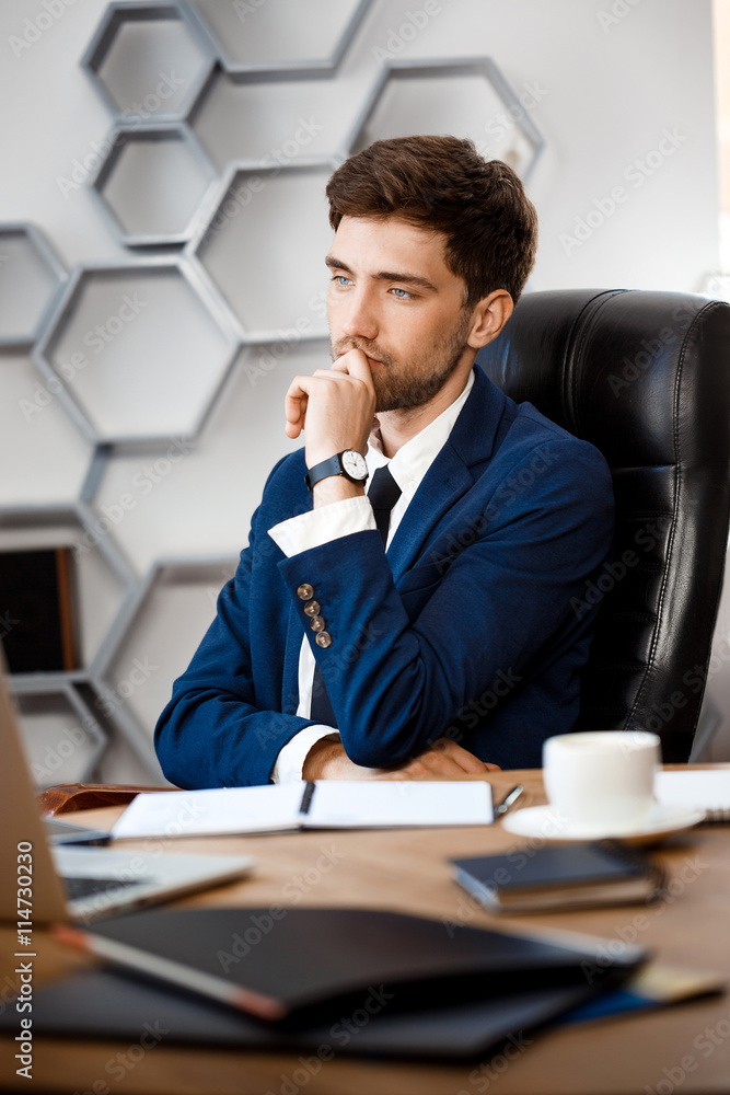 Young successful businessman sitting at workplace, office background. Young successful businessman sitting at workplace, office background.