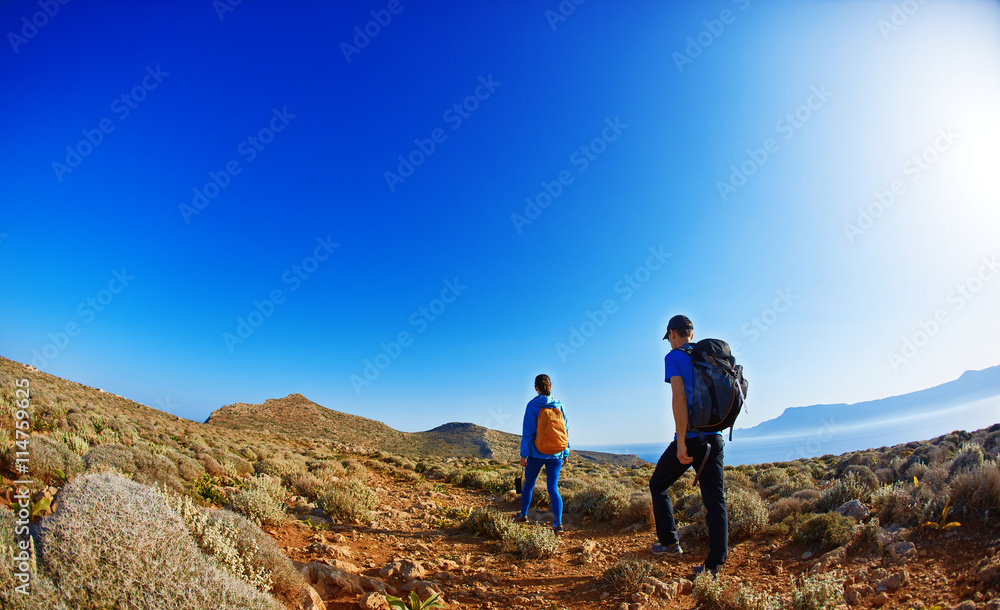 couple of travelers with backpack walking on the trail against sea and blue sky at early morning. Balos beach on background, Crete, Greece