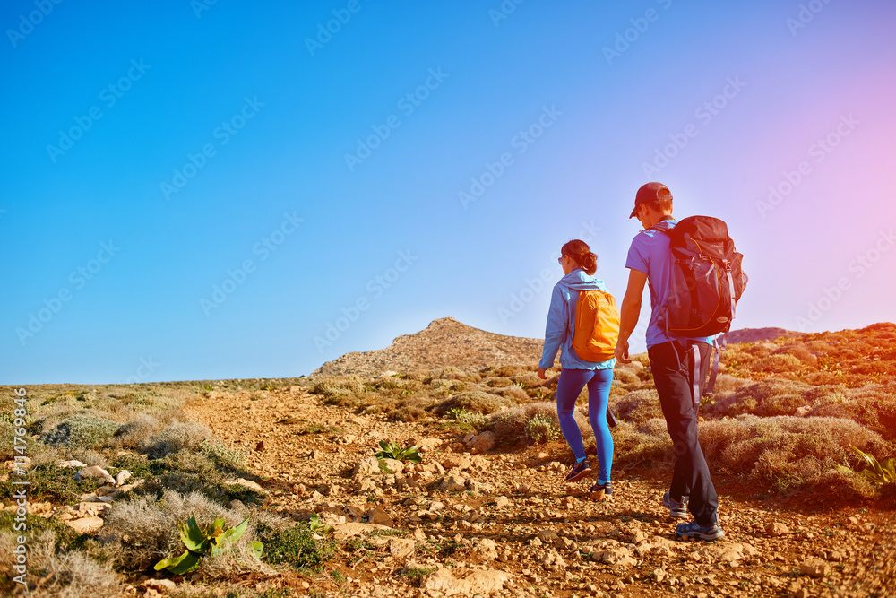 couple of travelers with backpack walking on the mountain trail against sea and blue sky at early morning. Balos beach on background, Crete, Greece