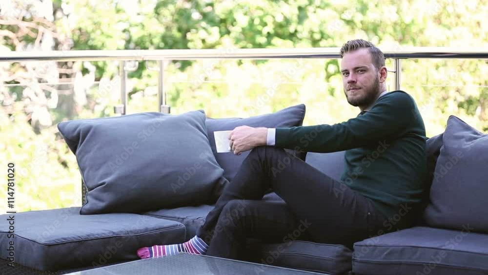 Man wearing a green pullover sitting down outside in a lounge sofa drinking from a mug. Looking at camera on a sunny day. Man wearing a green pullover sitting down outside in a lounge sofa drinking from a mug. Looking at camera on a sunny day.