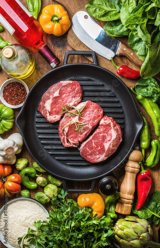 Ingredients for cooking healthy meat dinner. Raw uncooked beef steaks with vegetables, rice, herbs, spices and wine bottle over rustic wooden background,