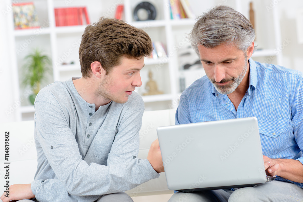 Father using laptop, teenage son next to him Father using laptop, teenage son next to him