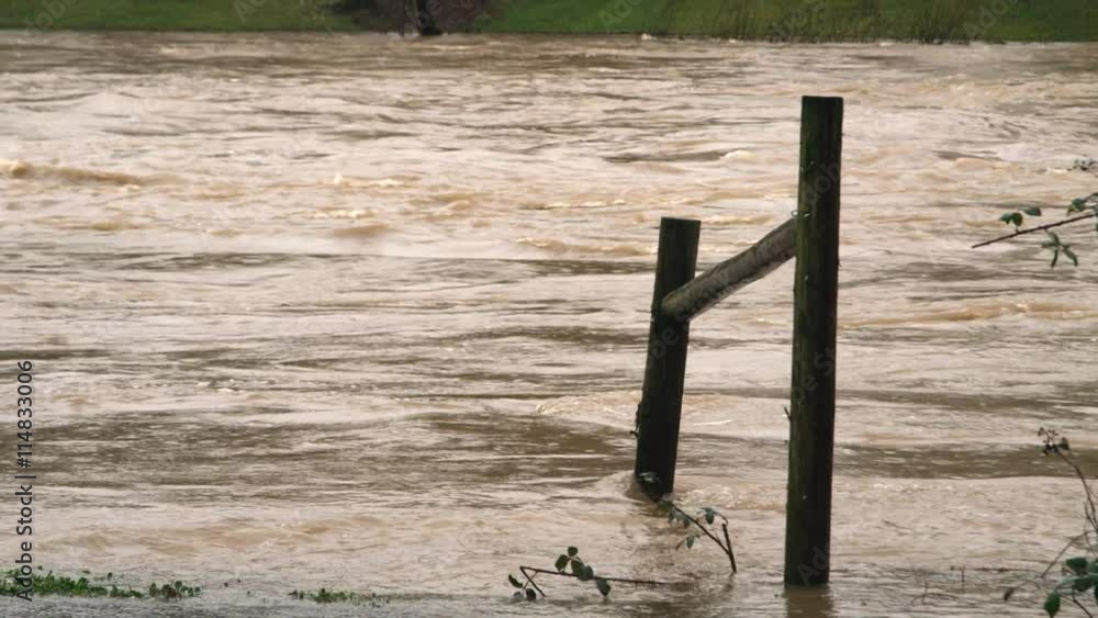 A swift muddy current washes around fence posts leading down to a flooding river A swift muddy current washes around fence posts leading down to a flooding river