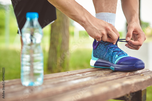 Drinking water from the plastic bottle in the park and running footwear close up. City outdoor workout and fitness healthy nutrition concept. Female athlete tying sport shoes laces before training.