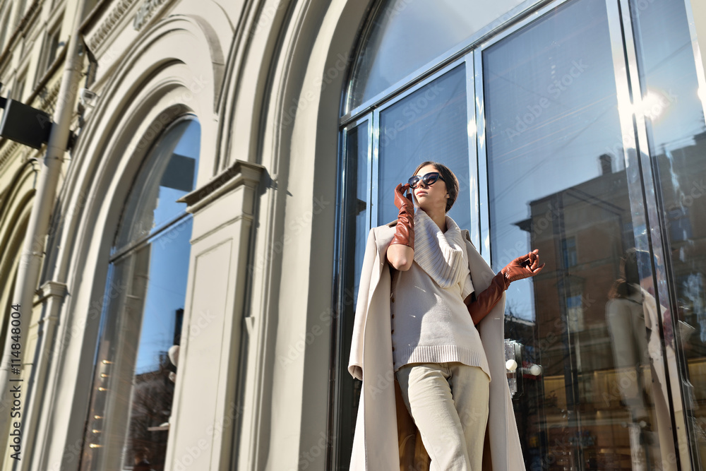 Fashionable portrait of lady with long hair in city. Image of brunette model on bright background. Young beautiful human in coat, gloves and sunglasses. Adult female person wears street style outdoors Fashionable portrait of lady with long hair in city. Image of brunette model on bright background. Young beautiful human in coat, gloves and sunglasses. Adult female person wears street style outdoors
