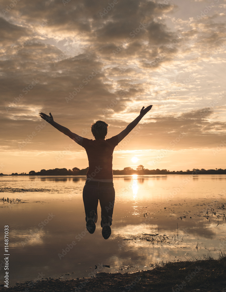 happy young slim woman jumping up on the shore of lake at sunset