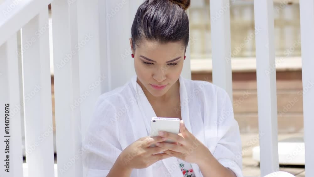 Attractive young woman with tied back hair sitting near fence smiling and texting with her smart phone outdoors Attractive young woman with tied back hair sitting near fence smiling and texting with her smart phone outdoors