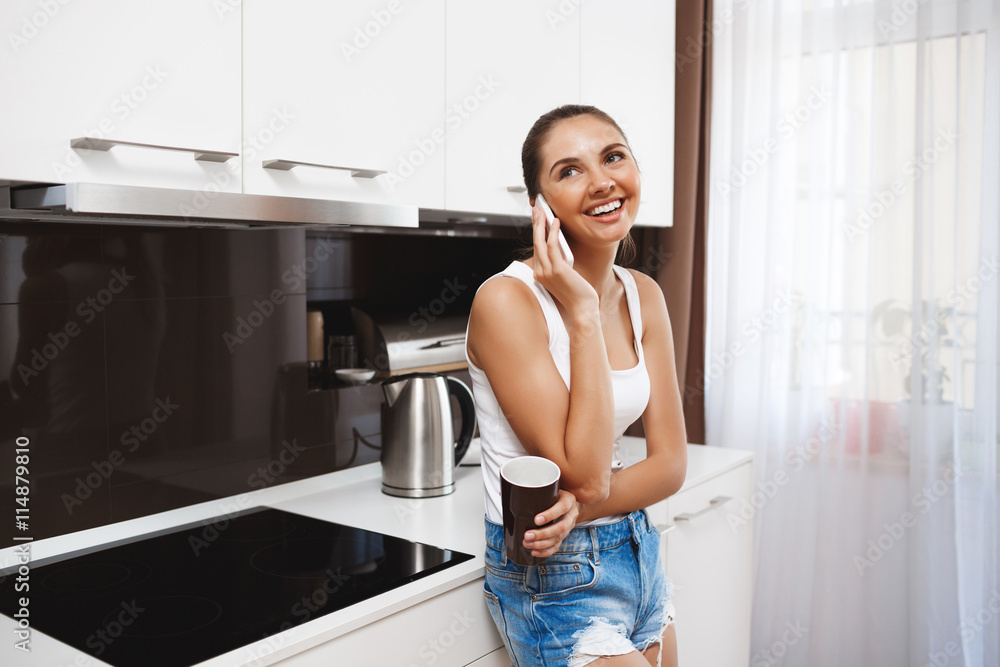 Beautiful smiling young girl talking on phone at kitchen and drinking a coffee Beautiful smiling young girl talking on phone at kitchen and drinking a coffee