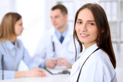 Young beautiful female doctor smiling on the background with patient and his doctor in hospital. High level and quality medical service concept.