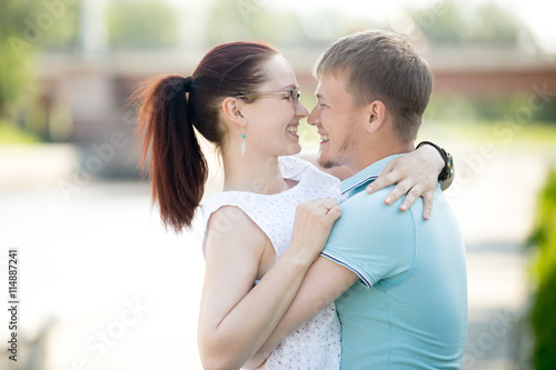 Beautiful happy young couple walking in summer city embracing and smiling at each other. Dark-haired glasses woman wearing white dress  looking at her handsome partner. People in love having fun