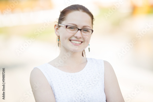Free Download Headshot portrait of cute pretty young woman wearing white dress and glasses in street and looking at camera with gentle smile against sunny street background - GFXLoad