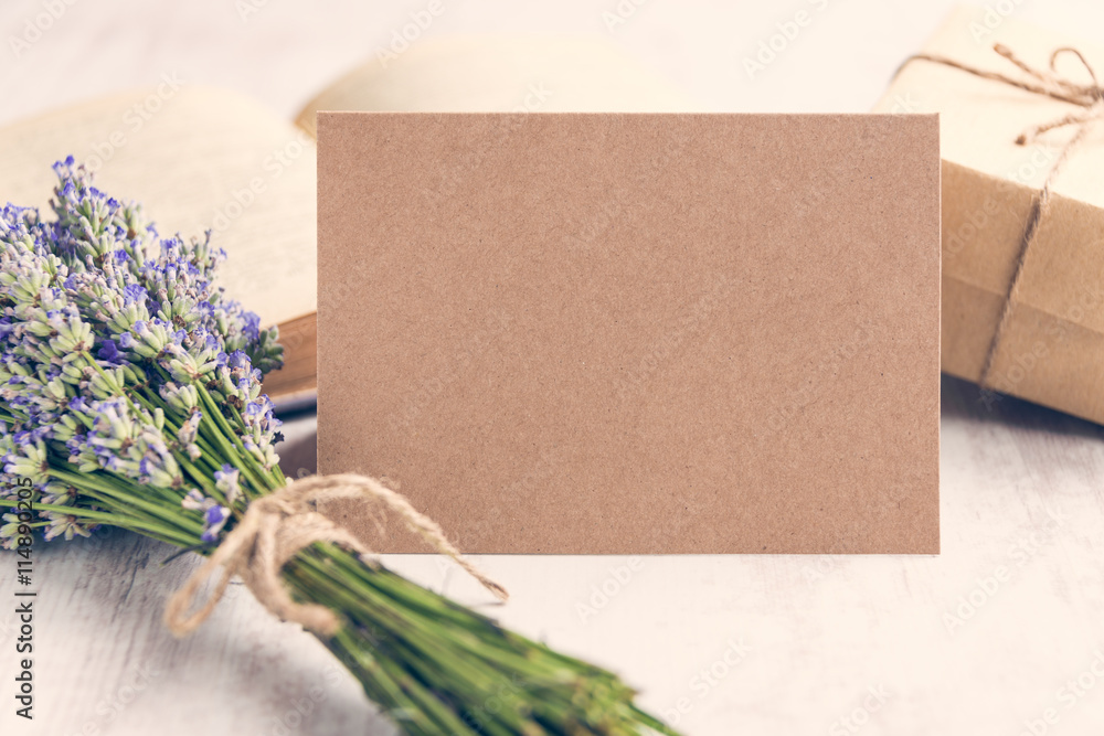 Empty greeting kraft card in front of a lavender bouquet, wrapped gift and old book over a white wood background. Vintage style.