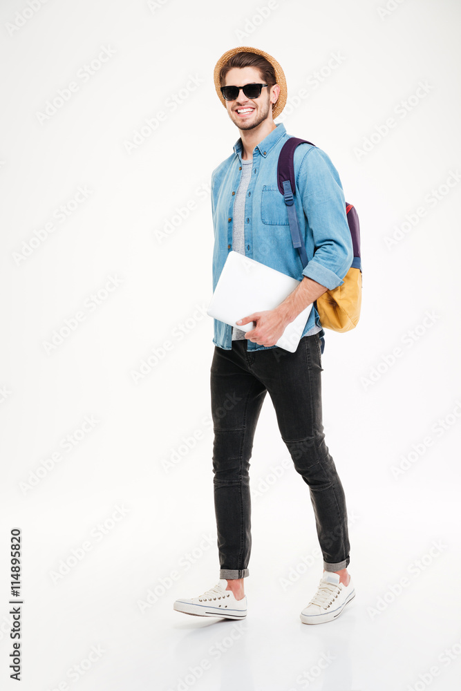 Fototapeta premium Cheerful young man with backpack walking and holding laptop