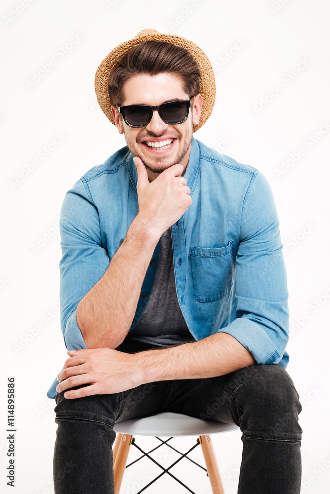 Smiling young man in hat and sunglasses sitting on chair Smiling young man in hat and sunglasses sitting on chair