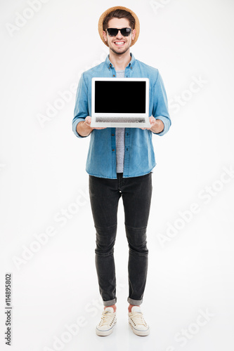 Happy man in sunglasses and hat holding blank screen laptop