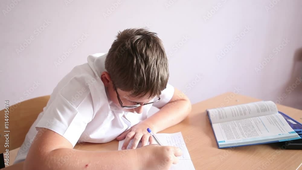 boy with glasses writing in a notebook sitting at a desk in the classroom. boy with glasses writing in a notebook sitting at a desk in the classroom.