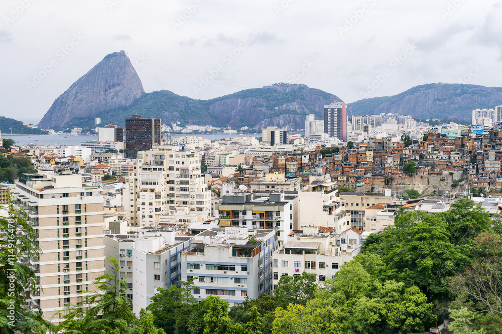 Gritty urban overlook of the Rio de Janeiro city skyline with Sugarloaf Mountain looming over residential apartments and a nearby favela adjacent to the hillside Santa Teresa neighborhood