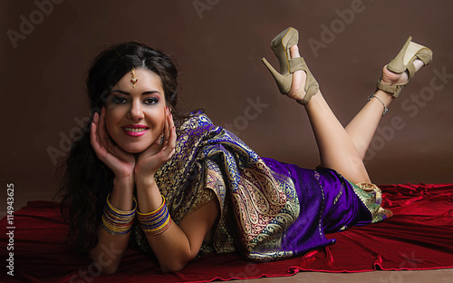 The young dark-haired woman in the rich Indian sari lying on bright red velvet and smiles placing hands under the face. Indian style.