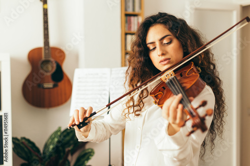 Girl playing violin. Young woman studying music alone at home in the living room with natural and soft light. Curly long and brunette hair, elegant dressed. photo