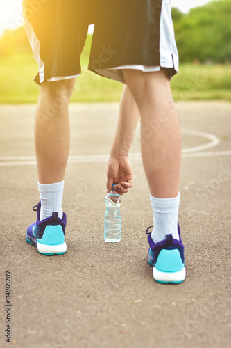 Drinking water from the plastic bottle in the park and running footwear close up. City outdoor workout and fitness healthy nutrition concept. Female athlete tying sport shoes laces before training.