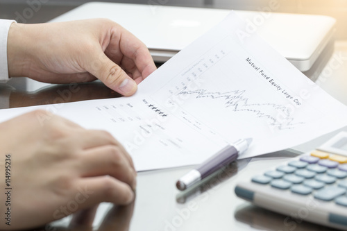 Businessman or fund manager review Mutual Fund information with performance line graph and benchmark with pen, calculator and computer laptop.