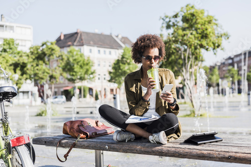 Young woman sitting on a bench drinking beverage while looking at her smartphone photo