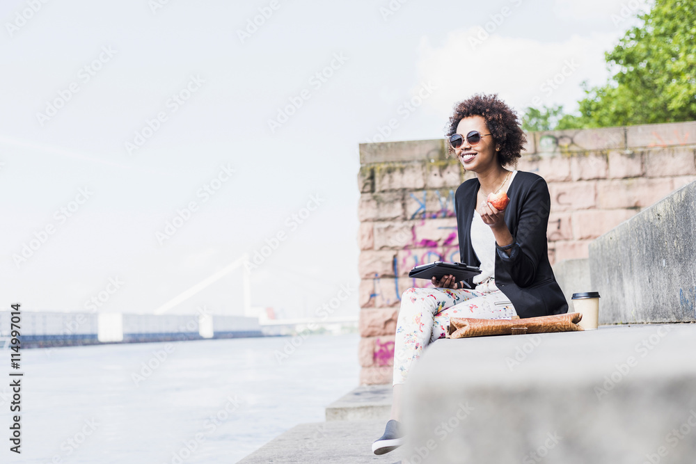 Germany, Mannheim, young woman with digital tablet eating an apple at riverside Germany, Mannheim, young woman with digital tablet eating an apple at riverside