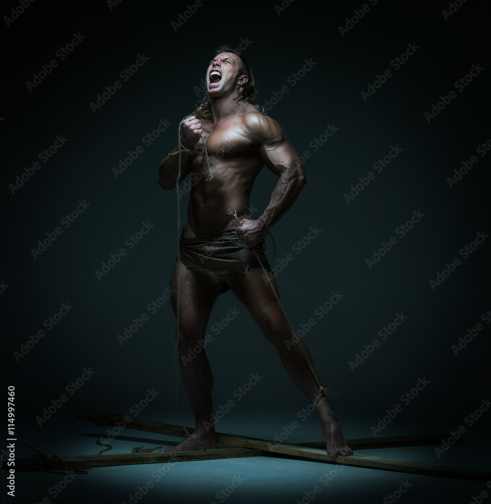 Screaming naked muscular man, hands tied rope to wooden beams Screaming naked muscular man, hands tied rope to wooden beams