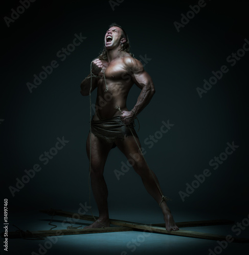 Screaming naked muscular man, hands tied rope to wooden beams