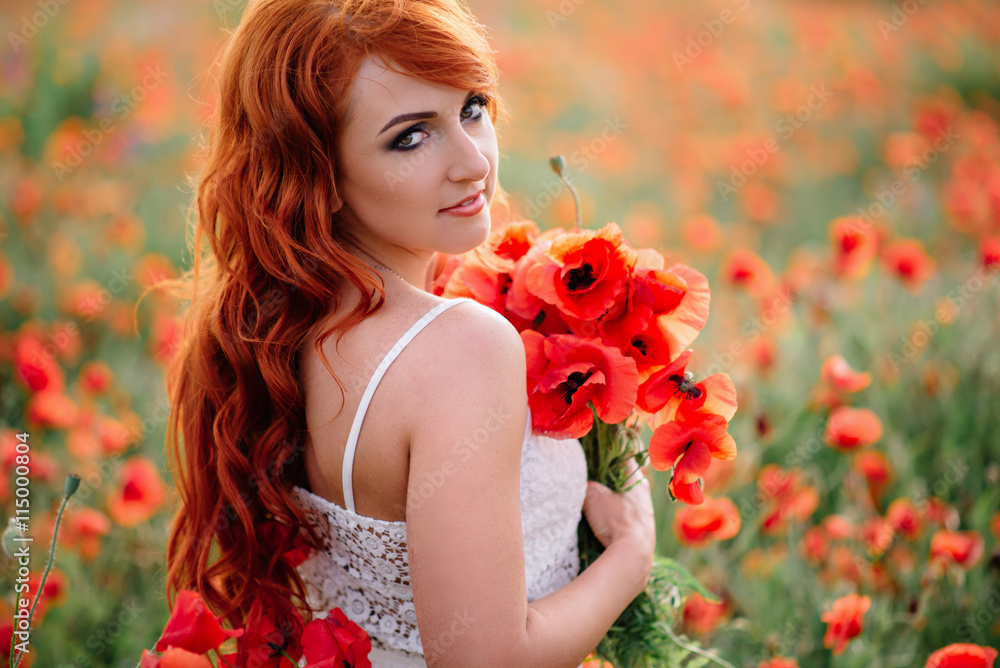 Naklejka premium beautiful young red-haired woman in poppy field holding a bouquet of poppies