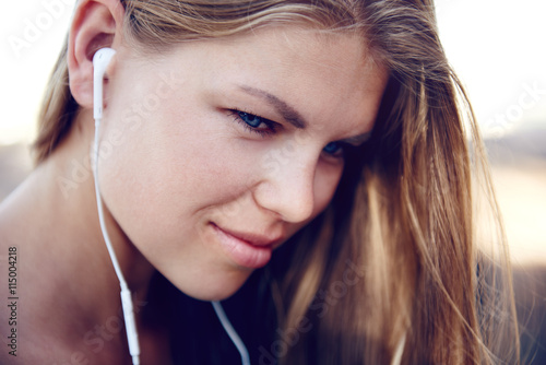 Close-up portrait of pretty female jogger with earphones listening music at dawn in the morning. Young fitness woman preparing to run outdoors. 