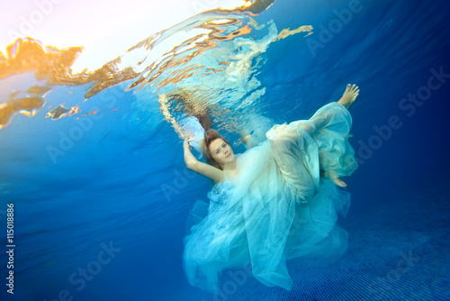 Beautiful girl dancing underwater in a white wedding dress against a blue background. Portrait. Horizontal orientation
