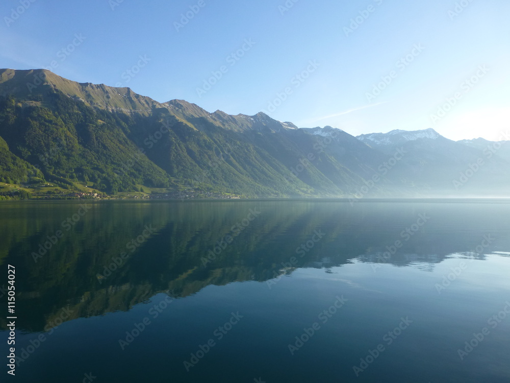 View across Brienzersee Lake, Switzerland on a sunny morning in late Spring, with the surrounding mountains reflected in the calm waters of the lake