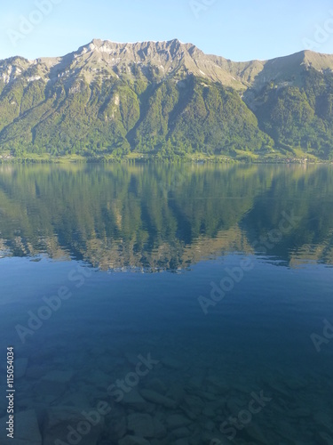 Early morning shot of Brienzersee lake, Switzerland, with mountain range opposite reflected in the calm lake, and submerged rocks visible through the clear water in the foreground