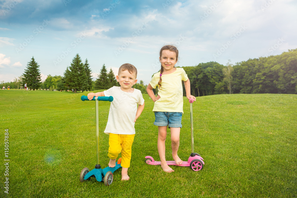 Fototapeta premium Two boy and a girl standing on the summer green field. Kick scooter riders. Shirts without prints templates you can place your picture or tex label brand