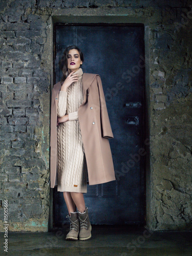 Elegant fashion model wearing long mocha coat, posing before a door.